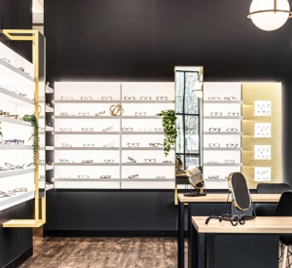Ein modernes Optikergeschäft mit schwarzen Wänden, beleuchteten Regalen mit verschiedenen Brillen, einer kleinen Pflanze, Spiegeln und Holztischen mit Brillen und Zubehör.