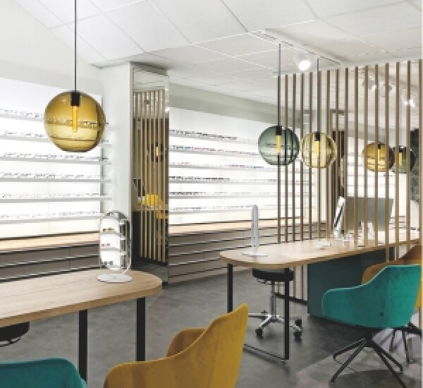 Modernes Optikerbüro mit Holzschreibtischen, tealfarbenen und gelben Stühlen, Glaspendelleuchten und Regalen mit vielen Brillen an einer weißen Wand im Hintergrund. Der Raum hat ein klares, zeitgenössisches Design.