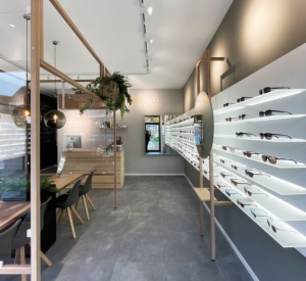 Modernes Optikergeschäft in schlichtem, minimalistischem Design mit gut beleuchteten Brillenregalen an der rechten Wand, einem Holztisch mit Stühlen auf der linken Seite und einer kleinen Theke im hinteren Bereich.