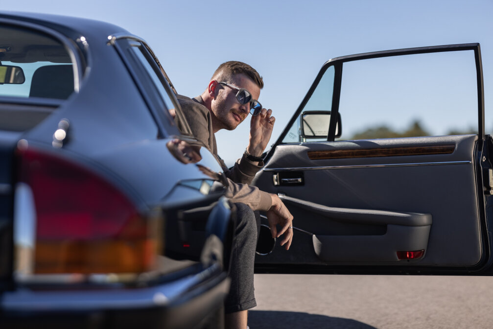 Ein Mann mit Sonnenbrille sitzt in einem Oldtimer mit offener Tür und schaut in die Kamera. Das glänzende Äußere des Autos und der klare Himmel lassen auf einen sonnigen Tag schließen.