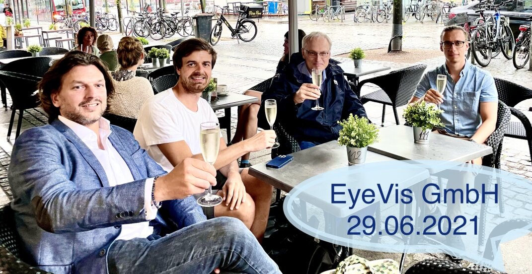 Vier M&auml;nner sitzen an einem Caf&eacute;tisch im Freien, l&auml;cheln und sto&szlig;en mit einem Glas Champagner an. Die Szene ist entspannt und leger. Ein blauer Text &uuml;berlagert EyeVis GmbH 29.06.2021. Im Hintergrund sind Fahrr&auml;der und Menschen zu sehen.