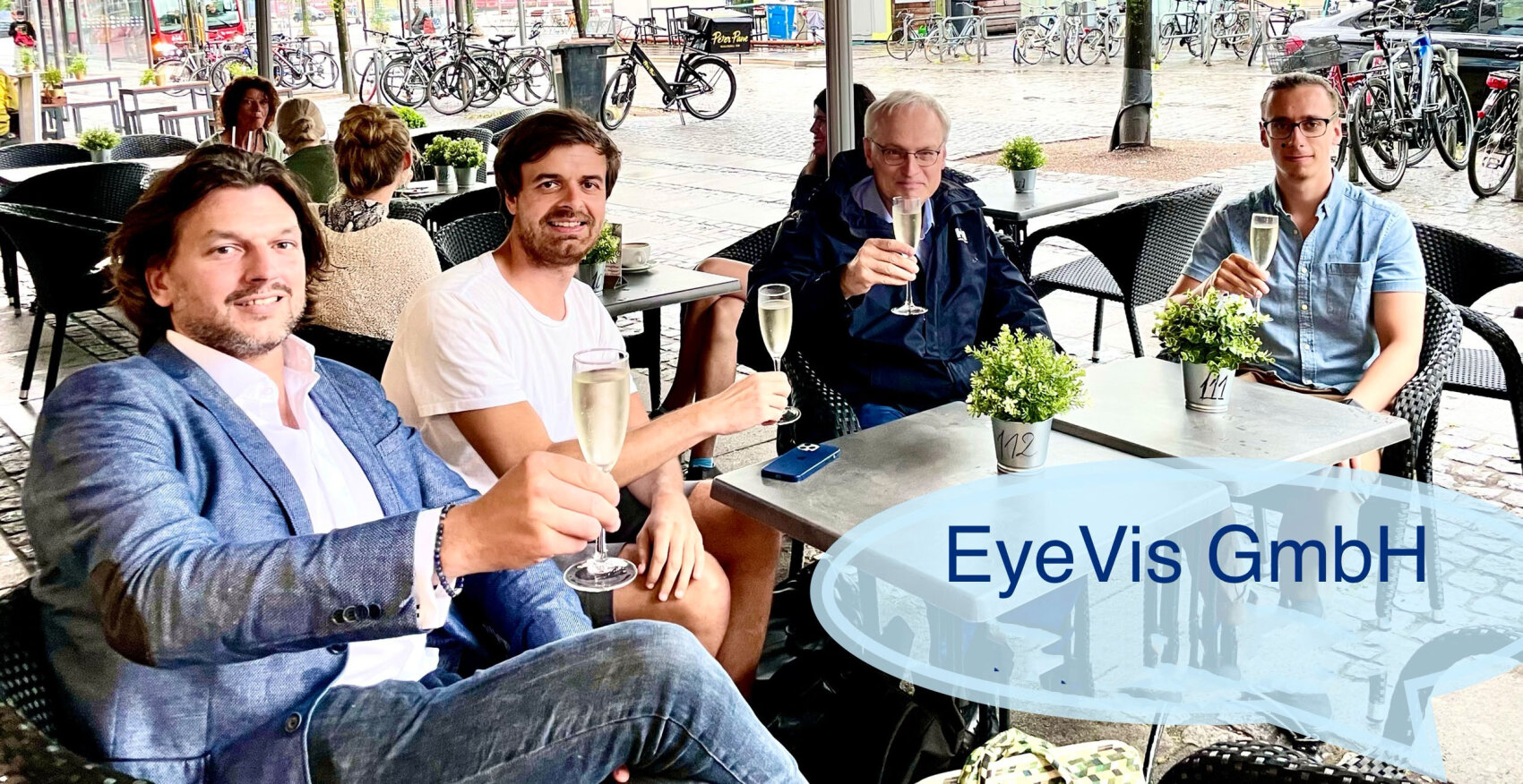 Four men sit at an outdoor café table, smiling and raising glasses in a toast. Small potted plants decorate the table. “EyeVis GmbH” text is on a transparent overlay in the bottom right corner. Bicycles are visible in the background.