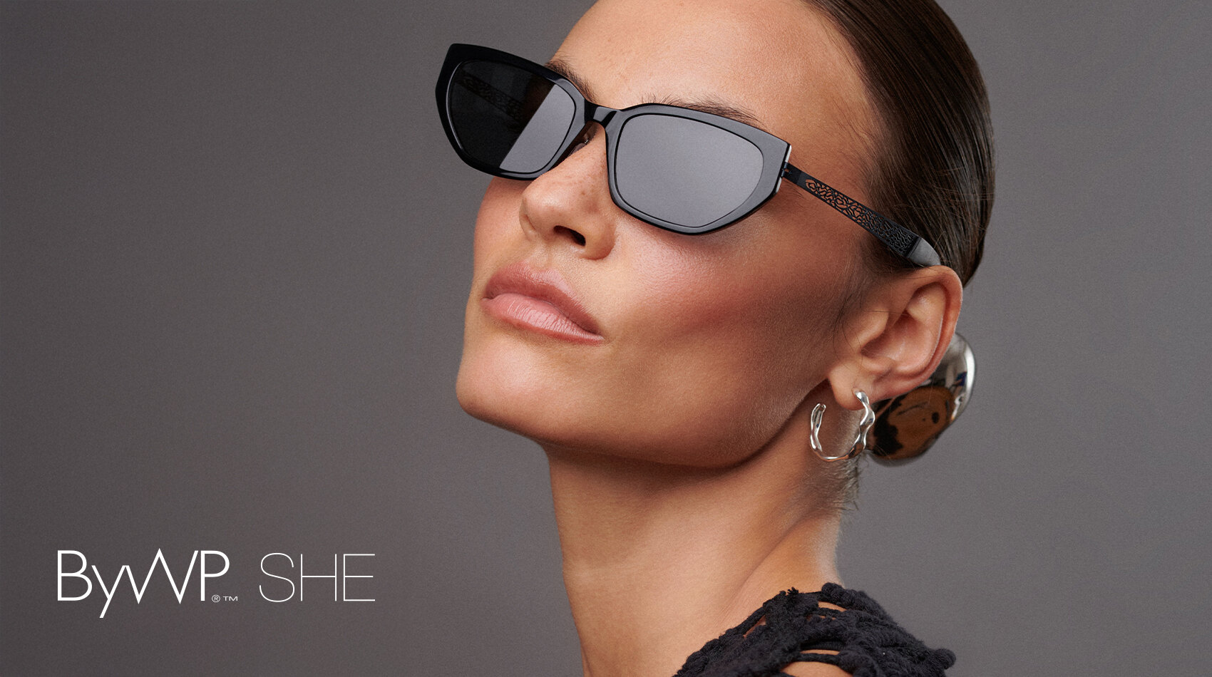 A woman with slicked-back hair wears black cat-eye sunglasses, earrings and a black textured top. The background is gray and the text ByWP SHE appears in the lower left corner.
