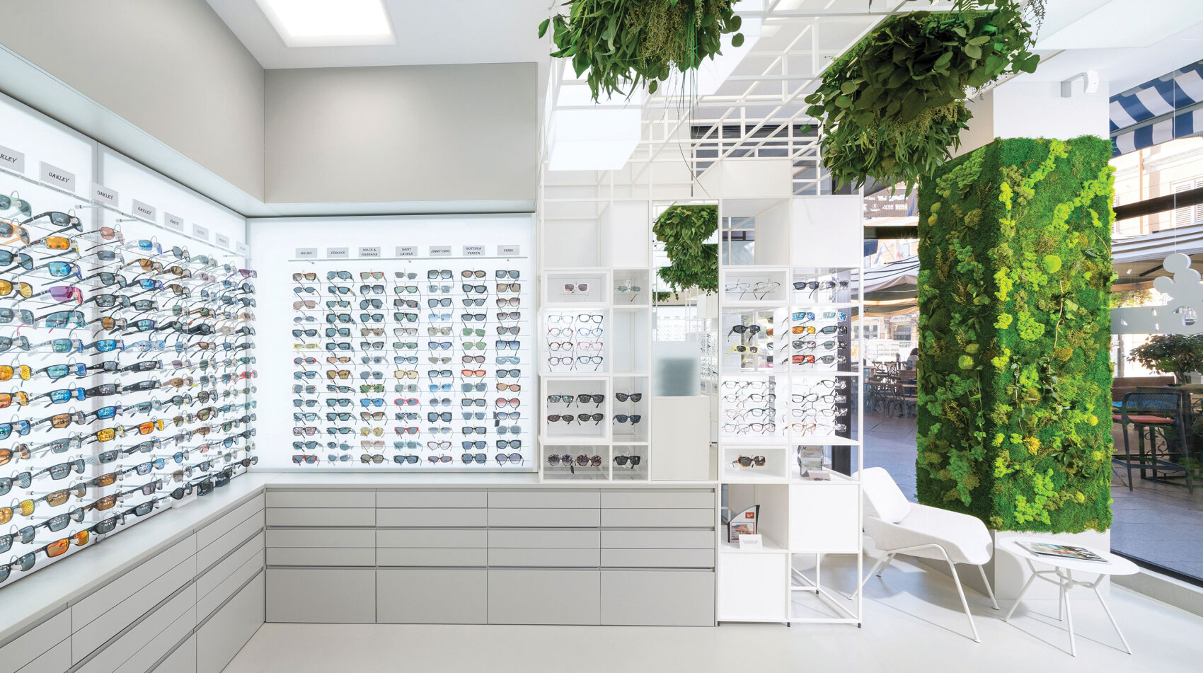 A modern eyewear store interior with white display shelves filled with eyeglasses and sunglasses, green vertical gardens, white chairs, and large windows letting in natural light.