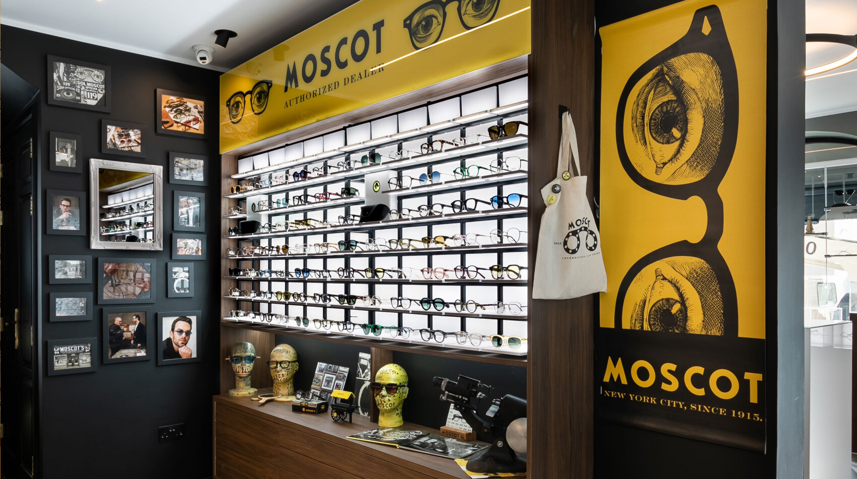 Ein stilvolles Brillengeschäft mit Brillenreihen, dem MOSCOT-Branding in Gelb und Schwarz, Postern im Vintage-Stil, gerahmten Fotos an einer dunklen Wand und Brillenaccessoires in Regalen.