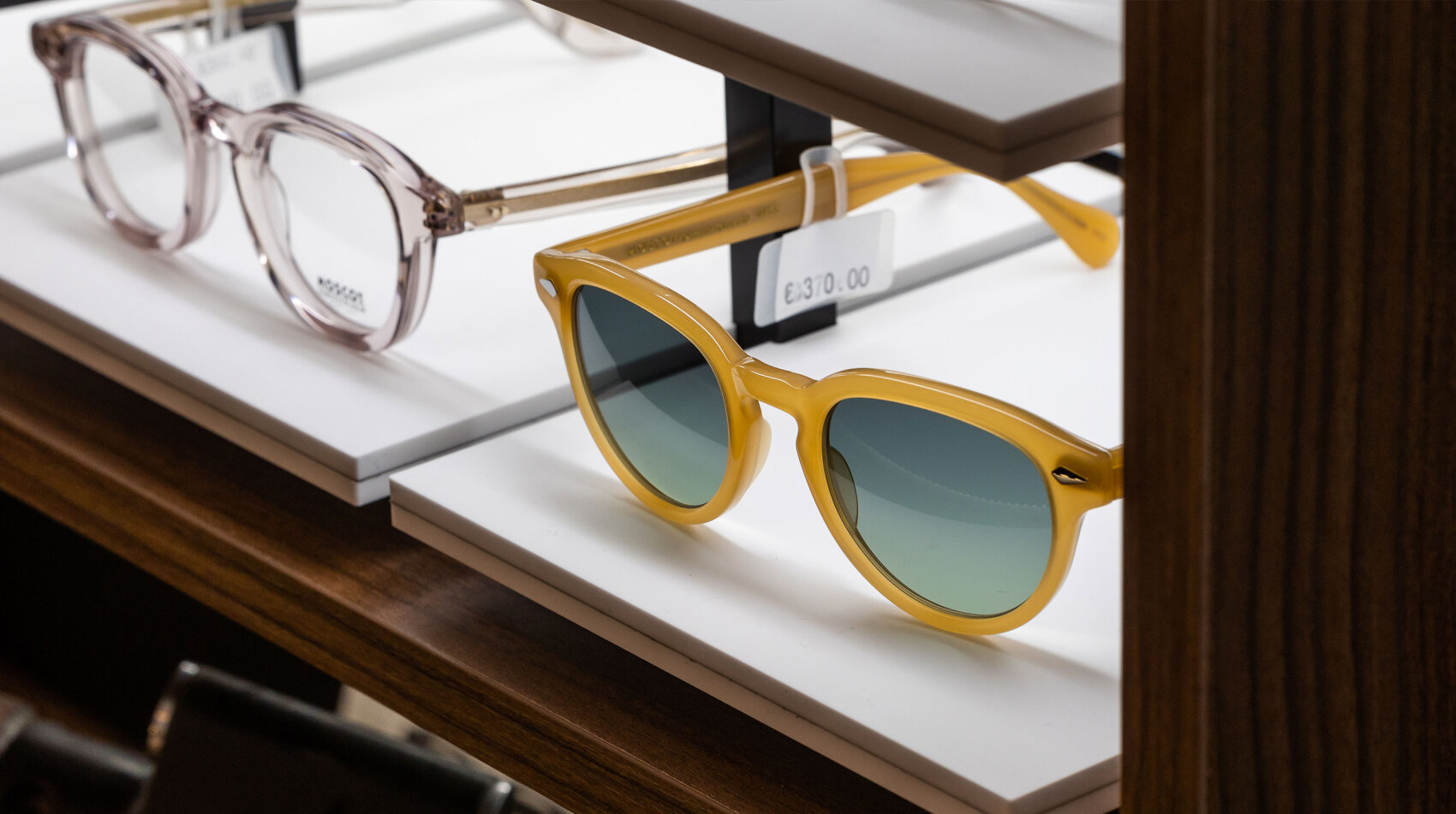 Eine gelbe Sonnenbrille und eine klare Brille werden in einem Geschäft in weißen Regalen ausgestellt. Neben der Sonnenbrille ist ein Preisschild mit der Angabe 130,00 € zu sehen.