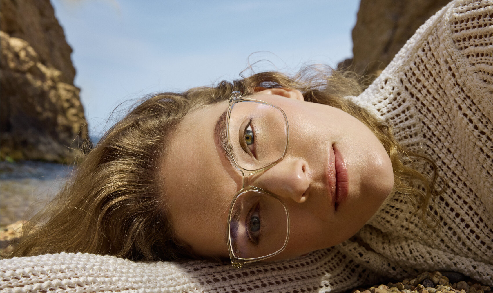 A person with wavy blonde hair and clear-frame glasses lies on a rocky beach, wearing a cream knit sweater, with cliffs and blue sky in the background.