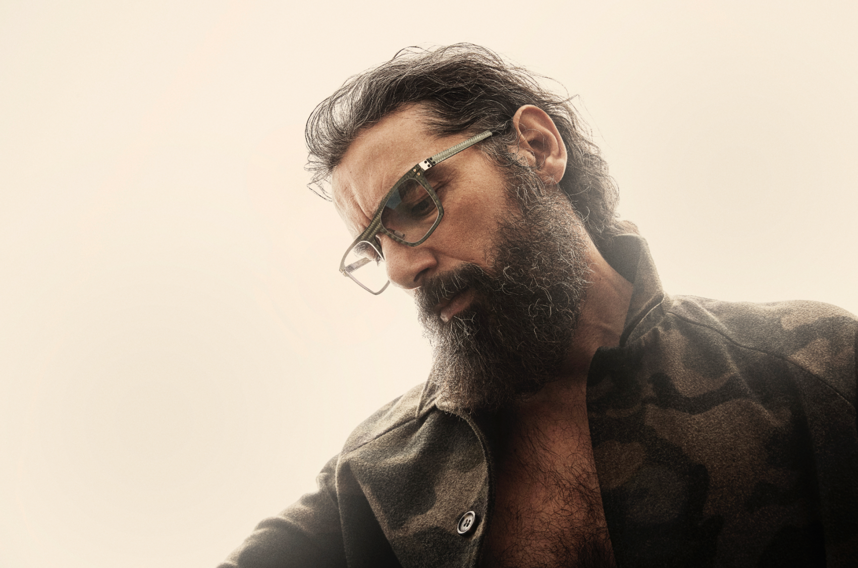 A bearded man wearing glasses and a camouflage-patterned jacket looks down thoughtfully against a light, blurred background.