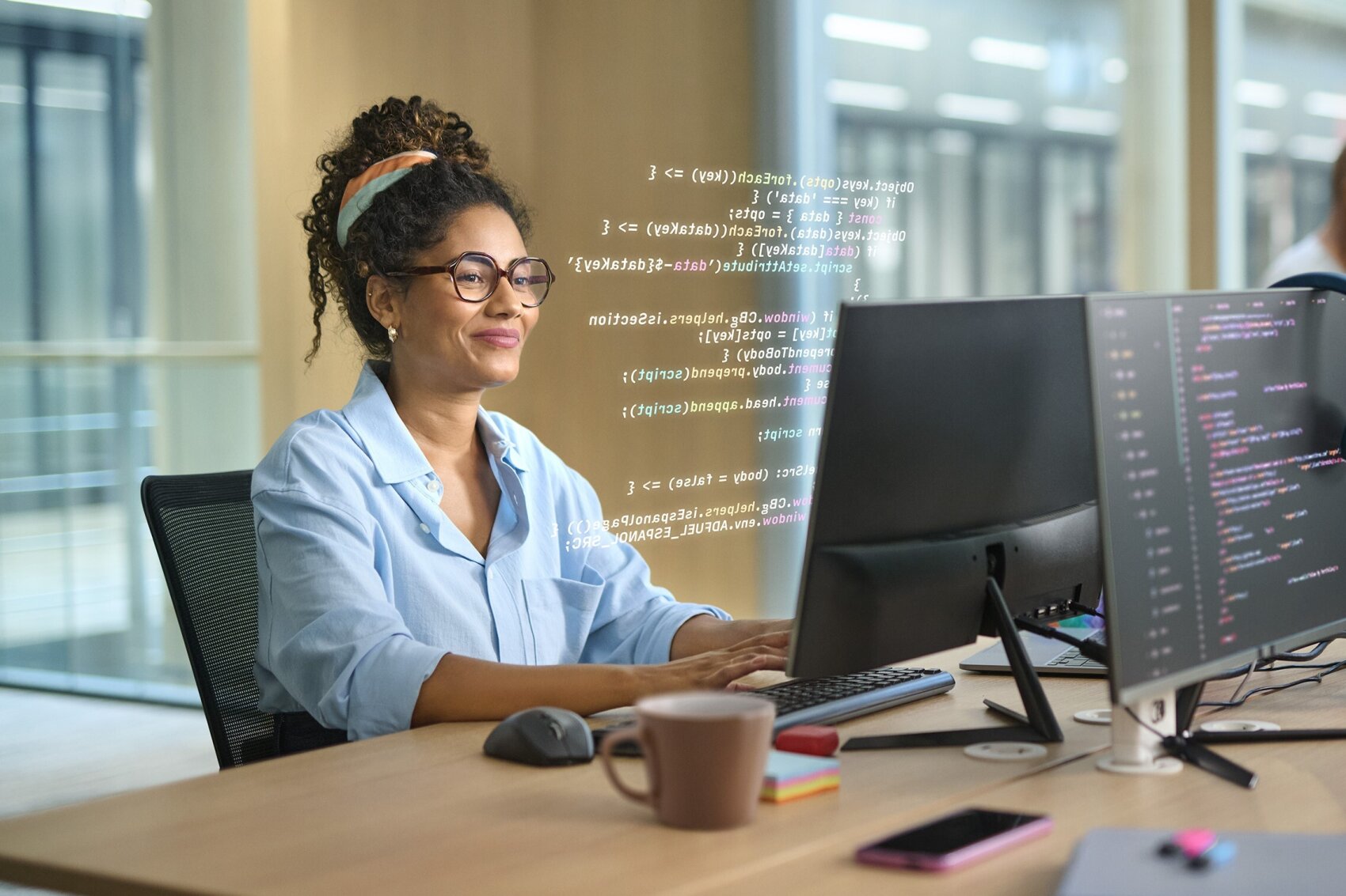 A woman wearing glasses sits at a desk with two monitors, typing on a keyboard. She is smiling, with programming code visible on the screens. A coffee mug and notepad are on the desk in a modern office setting.