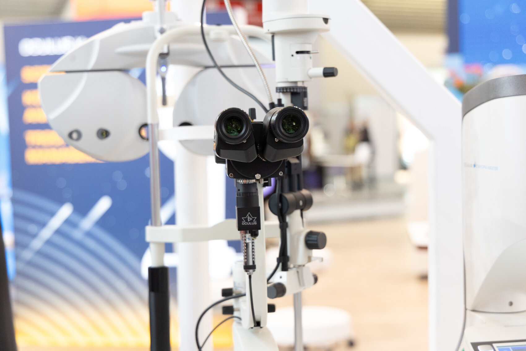 A close-up of an ophthalmic slit lamp microscope used for eye examinations, with blurred medical equipment and colorful background visible.