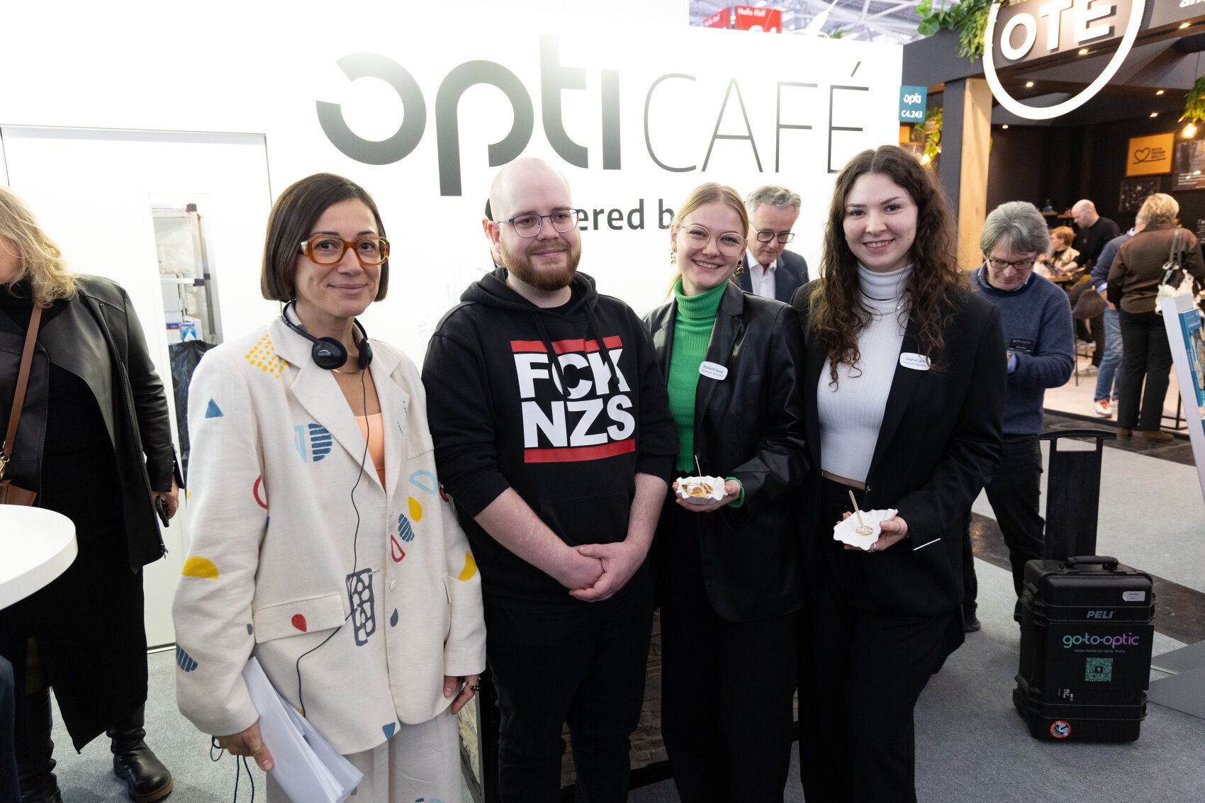 Four people stand together at an indoor event in front of a sign reading optICAFÉ. Three are smiling at the camera, two are holding food, and others are seen in the background. The atmosphere appears friendly and casual.